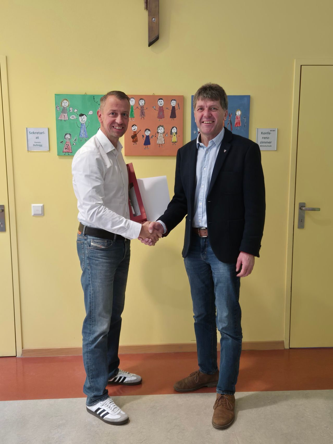 Zwei Männer schütteln sich die Hände in einem Raum mit gelben Wänden. Ein Mann hält eine Schachtel. Es gibt zwei Gemälde an der Wand. Eines ist mit Sekretariat beschriftet.