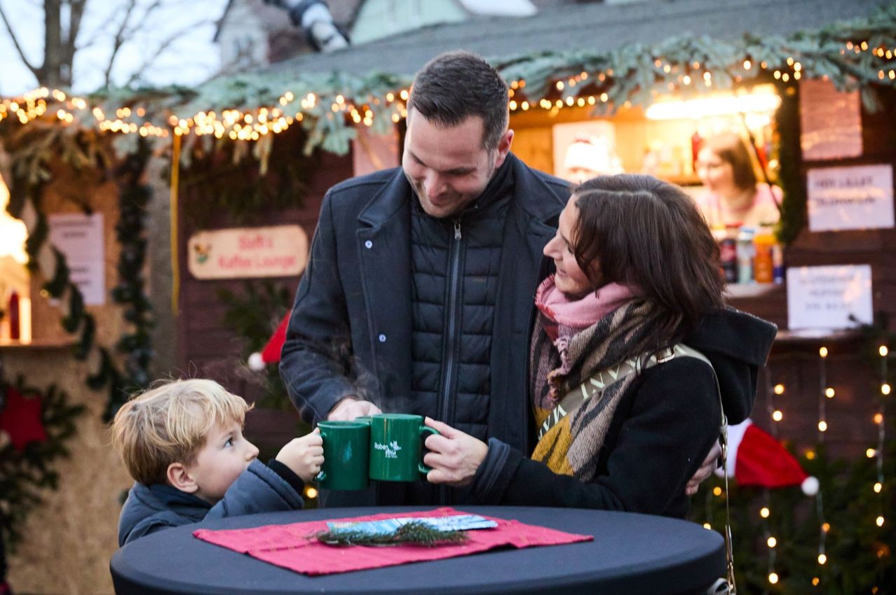 Eine dreiköpfige Familie genießt einen gemütlichen Moment gemeinsam auf einem Weihnachtsmarkt und teilt warme Getränke in festlichen Tassen.