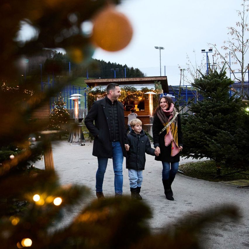 Eine dreiköpfige Familie läuft einen von Weihnachtsbäumen und Dekorationen gesäumten Weg entlang und lächelt für die Kamera.