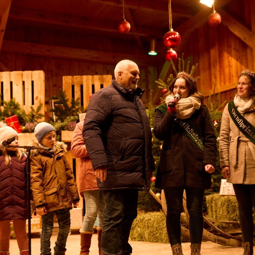 Ein Mann und eine Frau singen auf einer Bühne, während ein Mädchen ein Mikrofon hält, umgeben von Kindern in einer festlichen Umgebung mit Weihnachtsbäumen und Holzpaletten.
