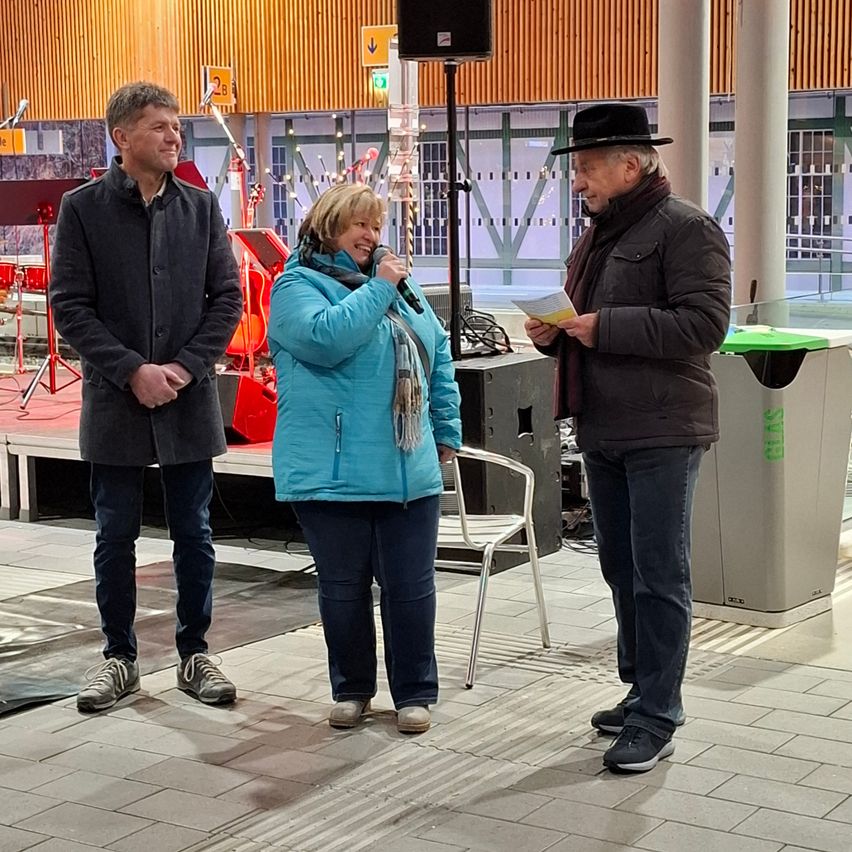 Drei Personen stehen vor einem Mikrofon. Die Frau hält ein Mikrofon, und der Mann auf der rechten Seite liest von einem Papier. Der andere Mann lächelt. Hinter ihnen ist eine Bühne mit Musikinstrumenten und einem Lautsprecher zu sehen.