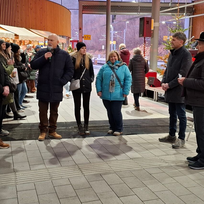 Eine Gruppe von Menschen ist versammelt, wahrscheinlich für eine Veranstaltung, mit einem Mann, der ein Mikrofon hält und spricht. Eine Frau in einem blauen Mantel lächelt, und andere hören zu. Hinter ihnen ist ein Weihnachtsbaum und ein Bus zu sehen.
