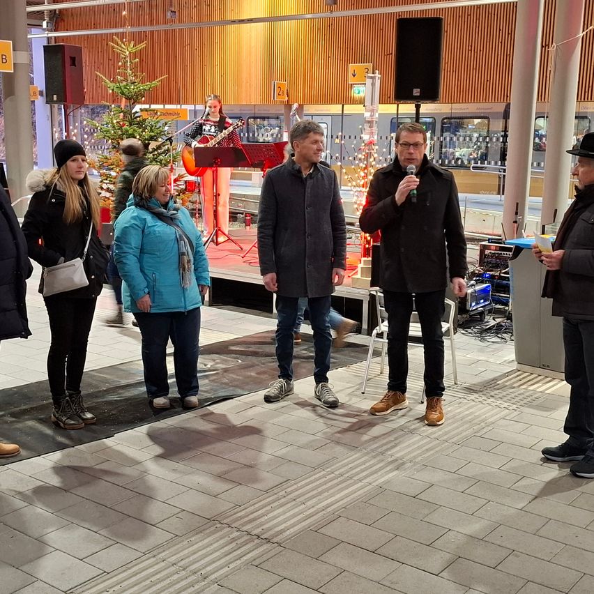Eine Gruppe von Menschen versammelt sich in einem Bahnhof. Einige stehen und andere gehen. Ein Mann spielt Gitarre auf einer Bühne. Ein Mann mit einem Mikrofon steht vor einem Lautsprecher. Ein Weihnachtsbaum und andere Dekorationen sind auf dem Bahnsteig platziert.