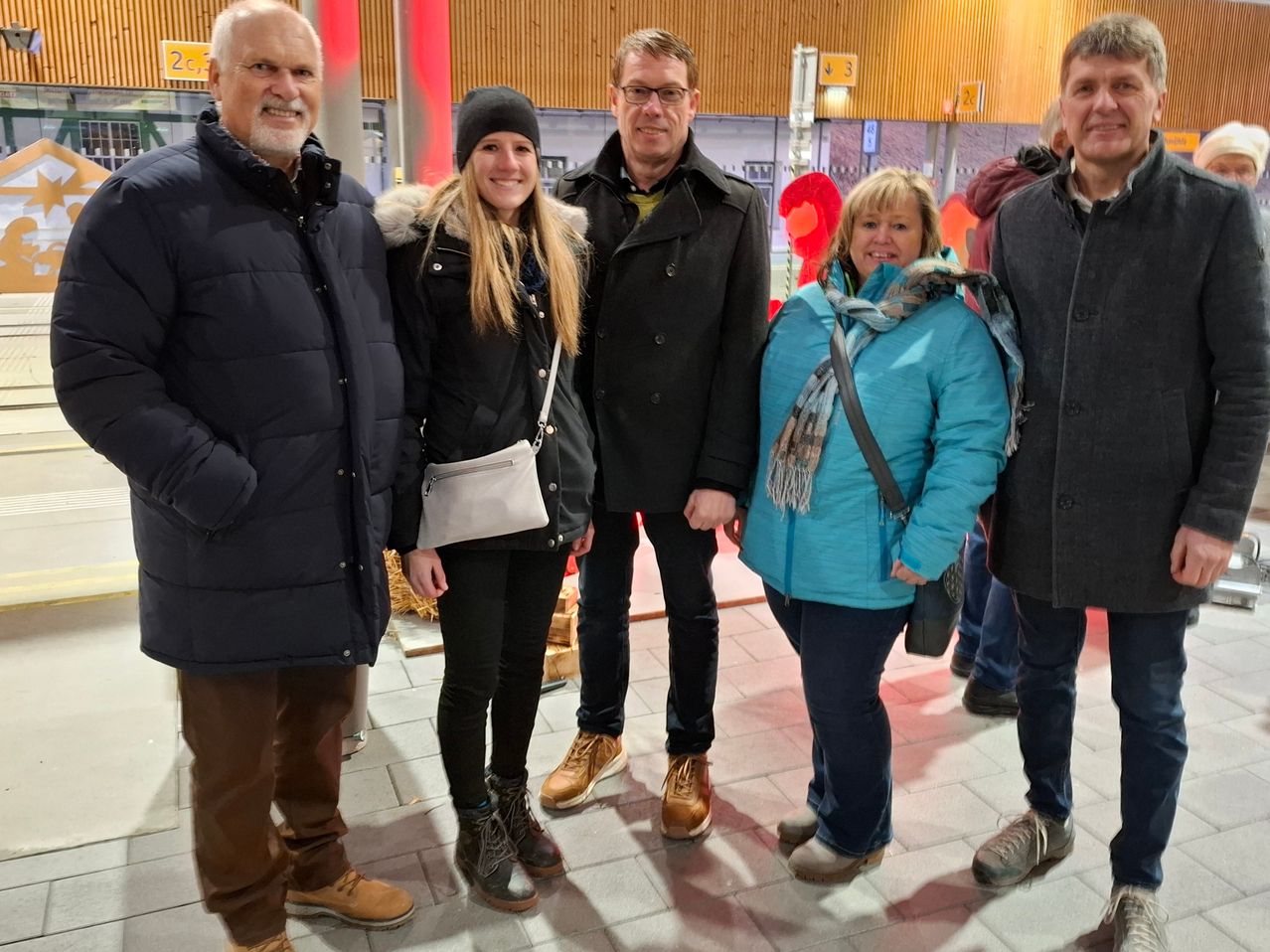 Fünf Menschen stehen zusammen auf einem gefliesten Boden und lächeln für ein Foto. Zwei Frauen und drei Männer, alle in Winterkleidung. Die Frau links hat eine weiße Tasche. Hinter ihnen ein Gebäude mit Schildern.