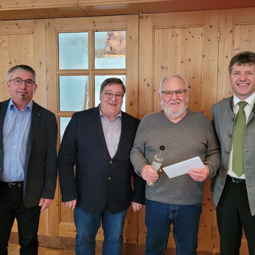 Vier Männer in formeller Kleidung stehen vor einer Holzwand. Der Mann in der Mitte hält eine Flasche und Papier. Der Mann auf der linken Seite trägt einen schwarzen Anzug, die anderen tragen graue und grüne Anzüge.