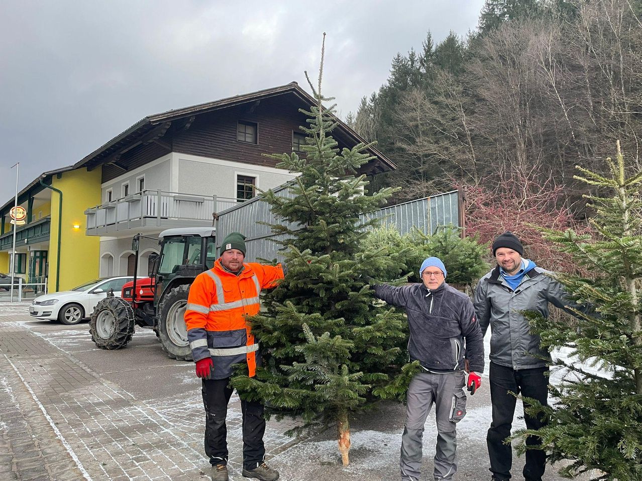 Drei Männer stehen vor einem Haus mit einem Traktor und Tannenbäumen. Ein Mann hält einen Baum. Der Boden ist verschneit.