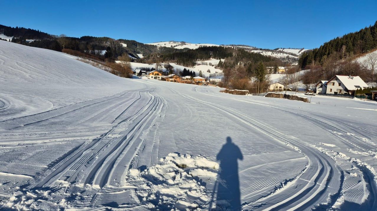 Eine verschneite Landschaft mit Skispuren auf dem Boden, einer Silhouette einer Person und mehreren Häusern im Hintergrund unter einem klaren blauen Himmel.