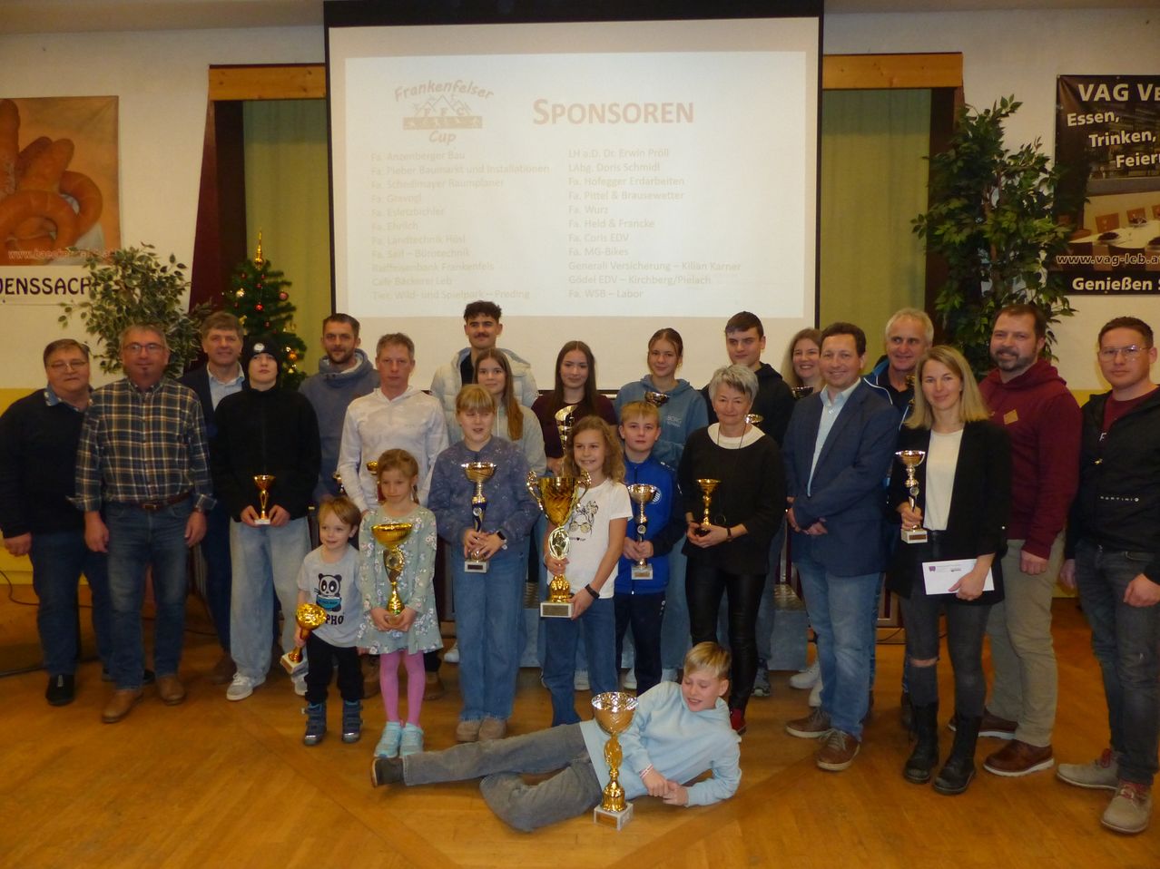 Eine Gruppe von Menschen in formeller Kleidung versammelt sich in einem Raum und hält Trophäen. Sie stehen vor einer Projektionsleinwand mit dem Text 'Sponsoren' und einem Weihnachtsbaum.