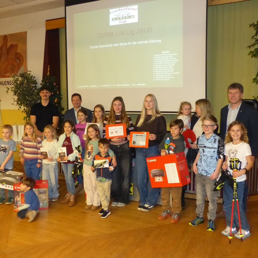 Eine Gruppe von Kindern, einige mit Urkunden in der Hand, steht vor einer Projektionsleinwand mit einer Präsentation. Dahinter stehen Erwachsene und verschiedene Gegenstände auf dem Boden.