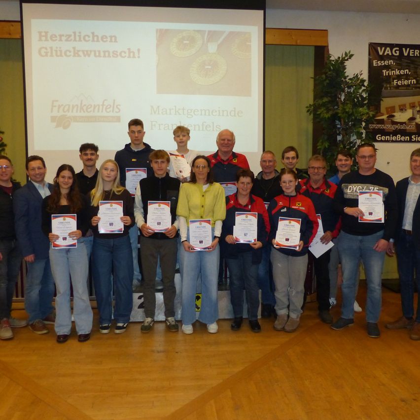 Eine Gruppe von Menschen posiert für ein Foto in einem Raum mit einer Projektionsleinwand, auf der 'Herzlichen Gluckwunsch!' steht und ein Zertifikat in der Hand.