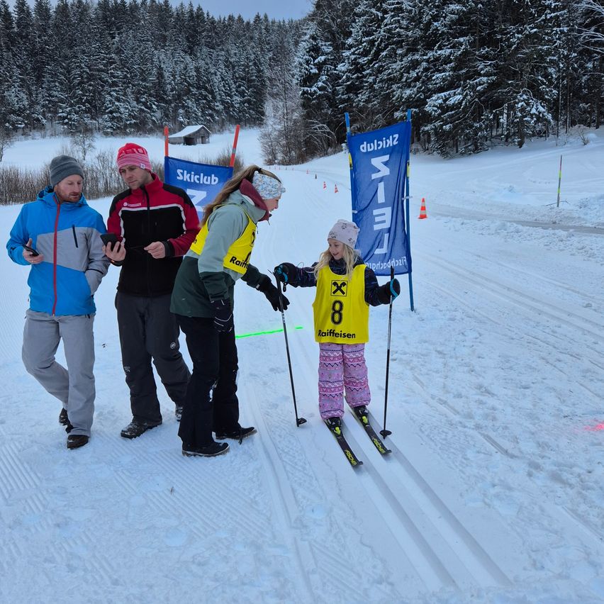 Ein Mädchen in einem gelben Nummer-8-Skianzug steht auf einem schneebedeckten Hang und wird von drei Erwachsenen in Skiausrüstung begleitet. Dahinter steht ein blaues Schild mit der Aufschrift 'Skiclub'.