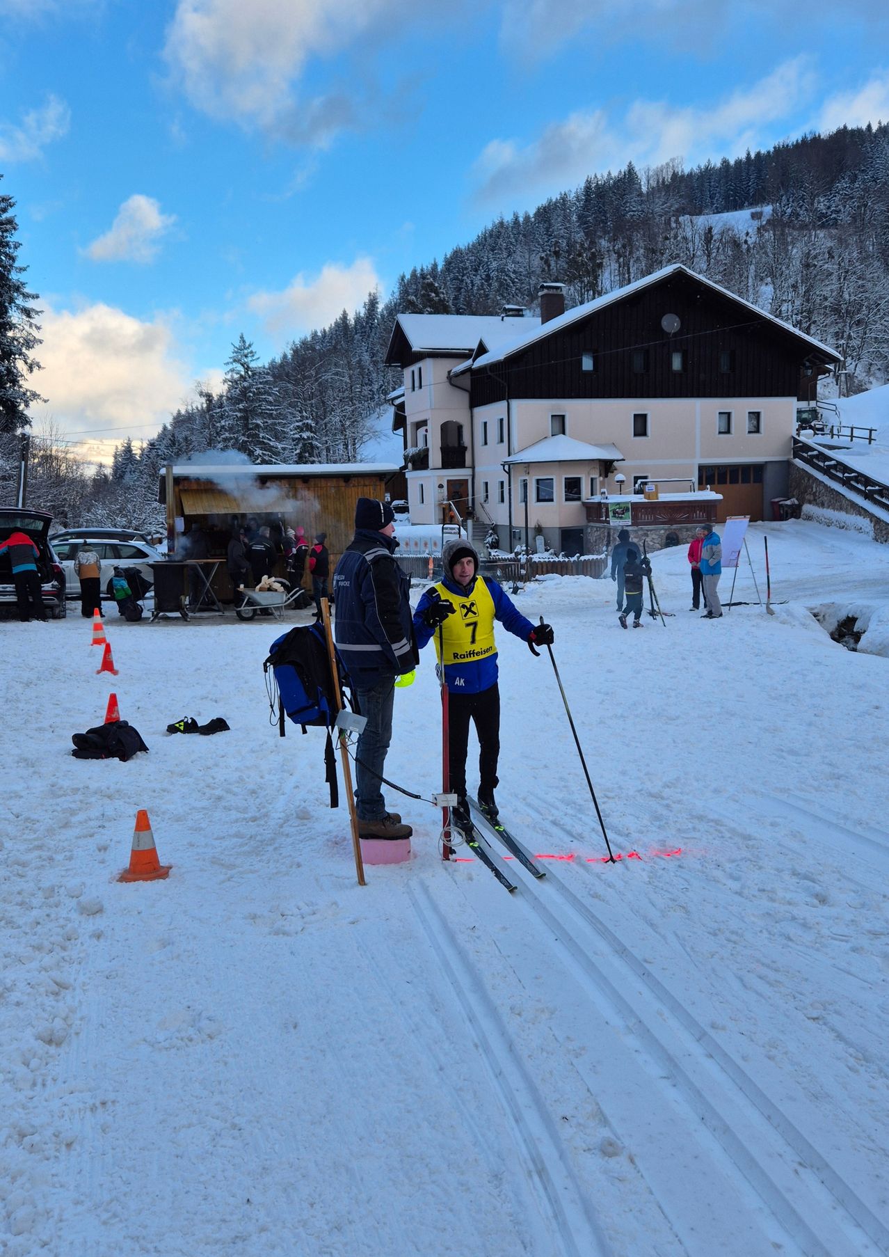 Ein Skifahrer mit gelber Weste steht auf einem verschneiten Pfad und bereitet sich auf ein Skirennen vor. Hinter ihm unterhält sich ein Mann in einer blauen Jacke. In der Nähe ein Haus und schneebedeckte Bäume.
