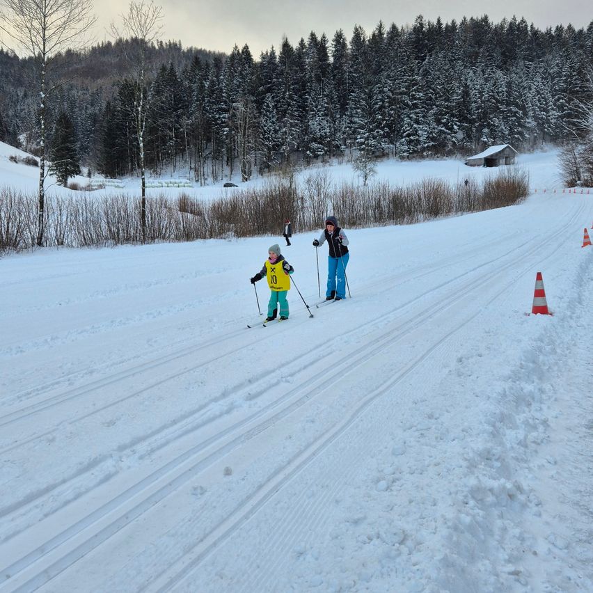 Zwei Personen langlaufen auf einem verschneiten Pfad. Eine Person trägt ein gelbes Jacket mit einer Nummer. Dahinter sind Bäume und ein Haus.