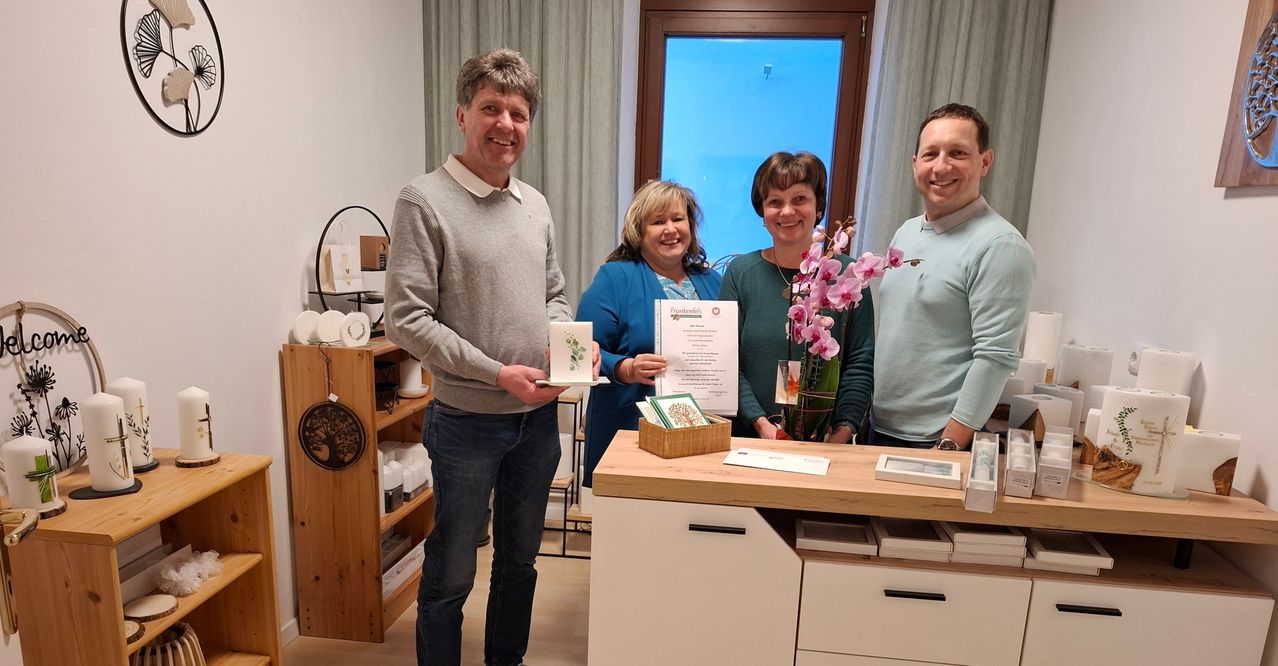 Vier Personen stehen in einem Raum, lächeln und halten ein Zertifikat und ein Buch. Eine Frau hält Blumen und ein Zertifikat, und eine andere Frau lächelt. Ein Schreibtisch mit Schubladen und ein Regal mit Gegenständen steht hinter ihnen.