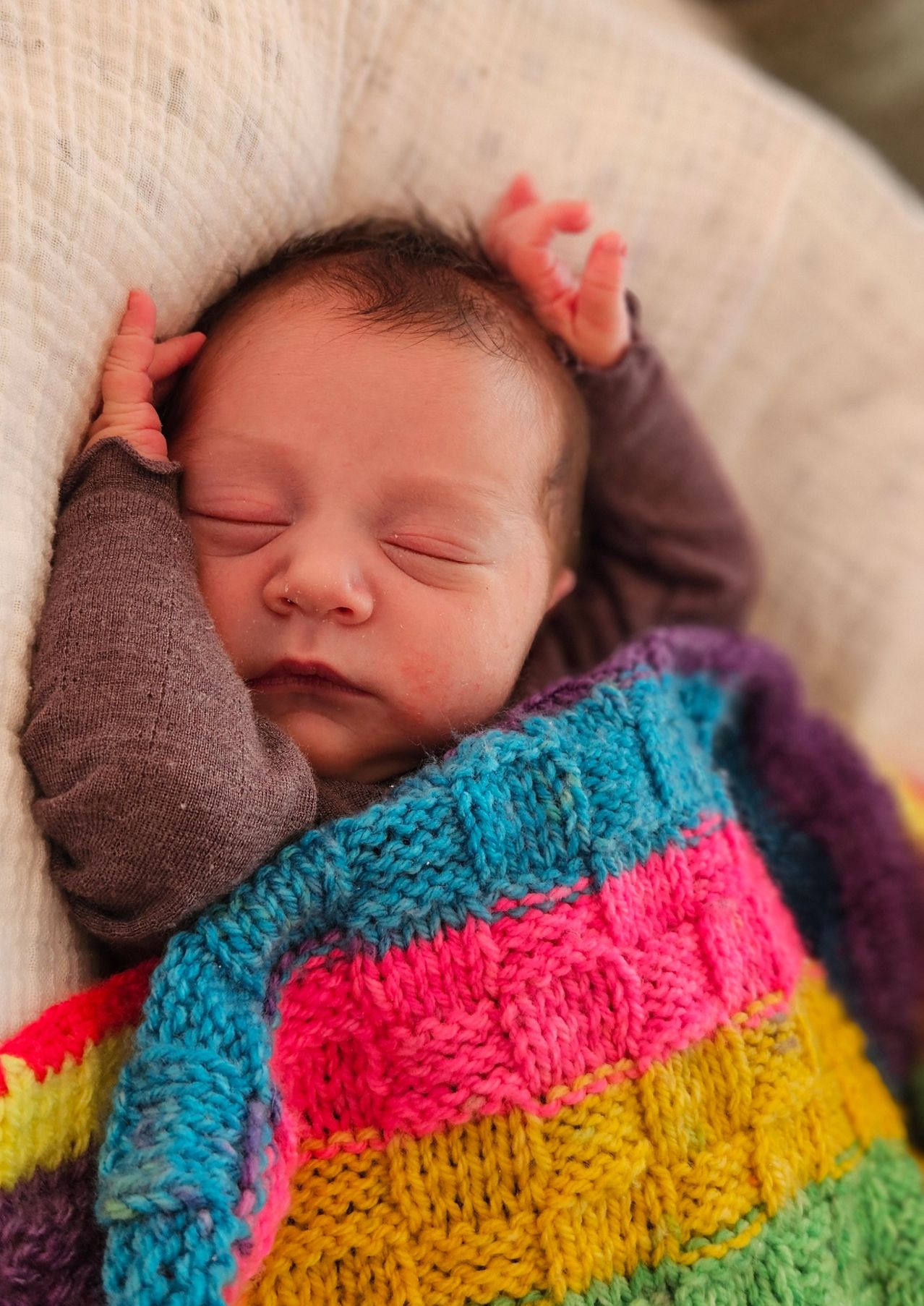 Ein neugeborenes Baby schläft friedlich, eingewickelt in eine bunte gestrickte Decke. Das Baby hat braune Haare und trägt ein langes braunes Oberteil.