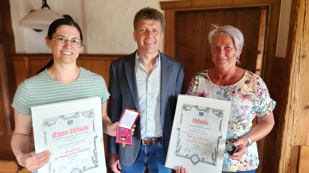 Drei Personen posieren mit Zertifikaten für ein Foto. Der Mann in der Mitte hält eine Medaillenschachtel. Eine Frau auf der rechten Seite hält ein Abzeichen. Die Frau auf der linken Seite hält ein Zertifikat.