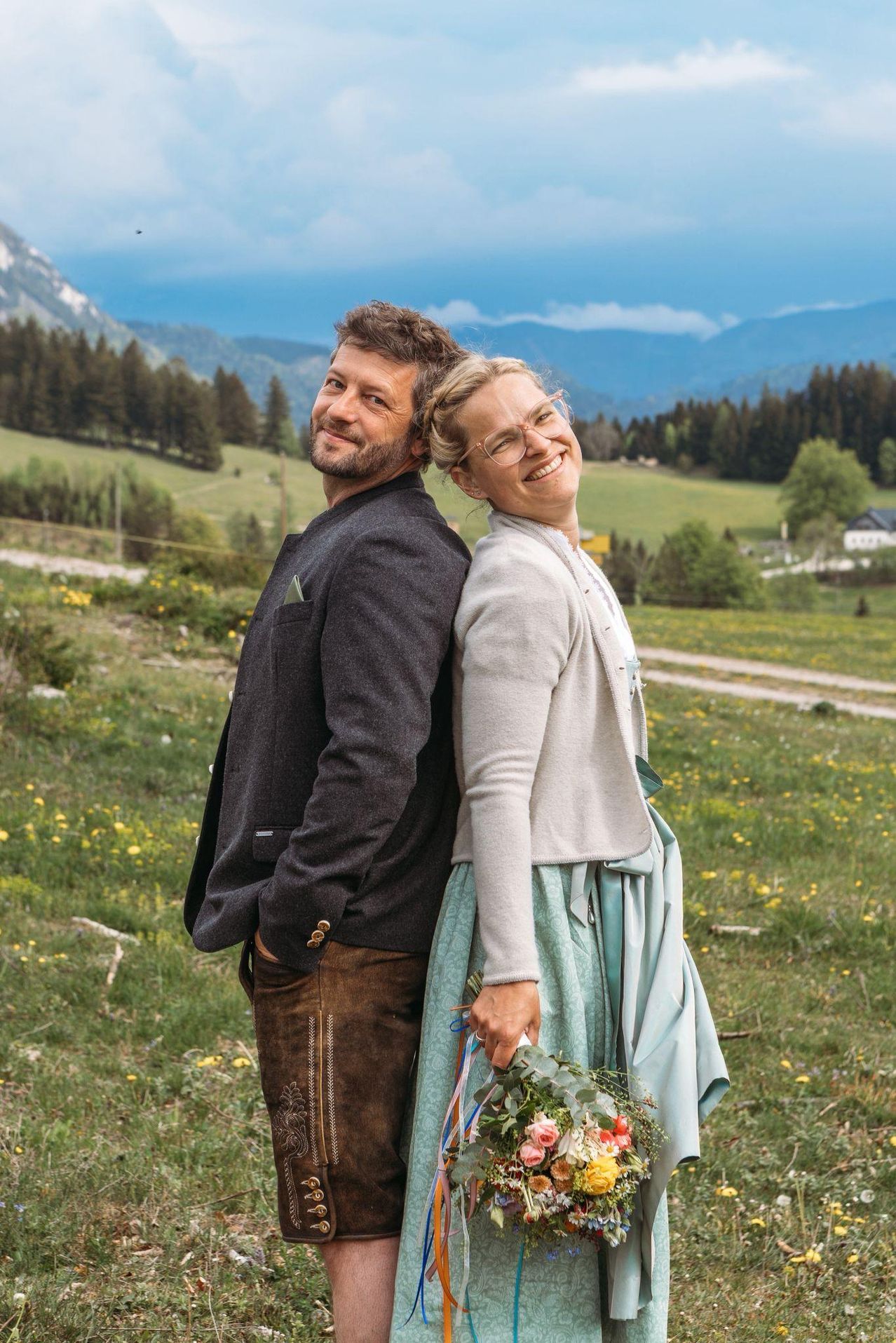 Ein Mann und eine Frau stehen in einem Grasfeld mit Bergen im Hintergrund und lächeln für ein Foto.