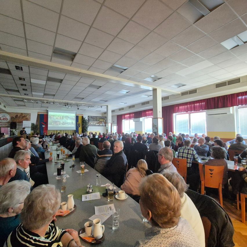 Ein großer Raum mit einer Projektionsleinwand vorne, viele Menschen sitzen an Tischen mit Tassen, Tellern und Papieren. Der Raum hat weiße Decken, rote Vorhänge und Glasfenster.
