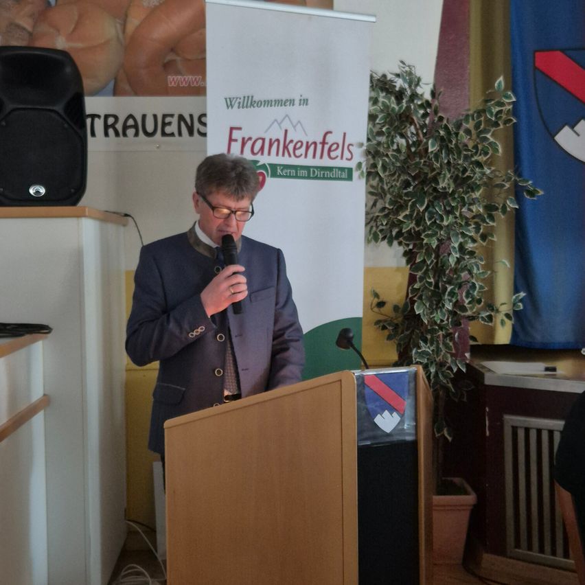 Ein Mann spricht in ein Mikrofon an einem Podium, mit einem Schild im Hintergrund, das 'Willkommen in Frankenfels' steht.