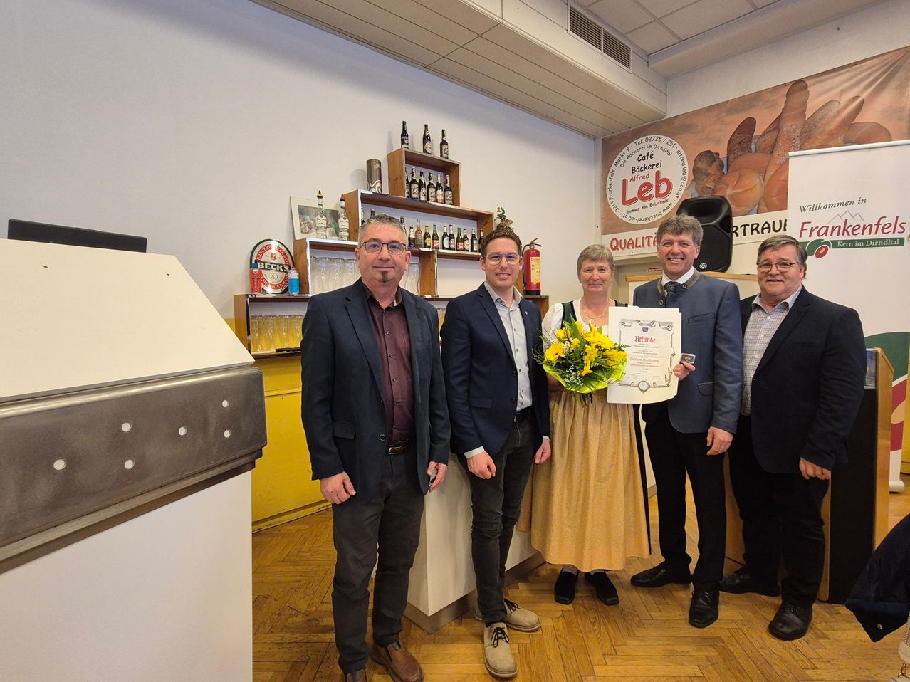 Fünf Personen posieren für ein Foto in einem Raum, einer hält ein Zertifikat und Blumen. Dahinter ist eine Wand mit einem Poster, einem Regal mit Flaschen und einem Schild.