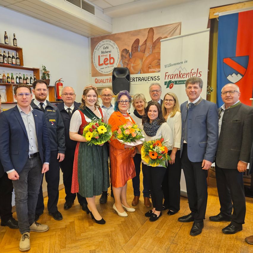 Eine Gruppe von Menschen in formeller Kleidung steht in einem Raum, einige halten Blumen. Ein Banner mit dem Text 'Willkommen in Frankenfels' ist an der Wand angebracht.