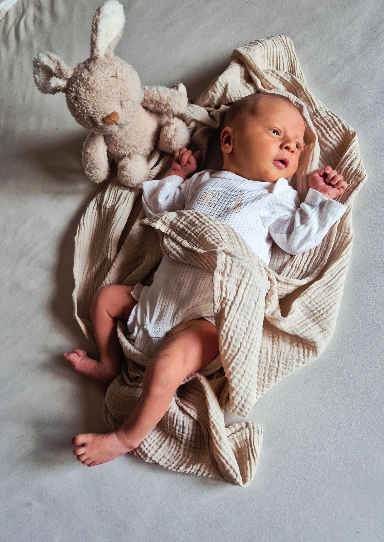 Ein Baby liegt auf einem Bett, in eine Decke gewickelt, schaut mit erhobenen Händen nach oben, neben einem beigefarbenen Teddybär.