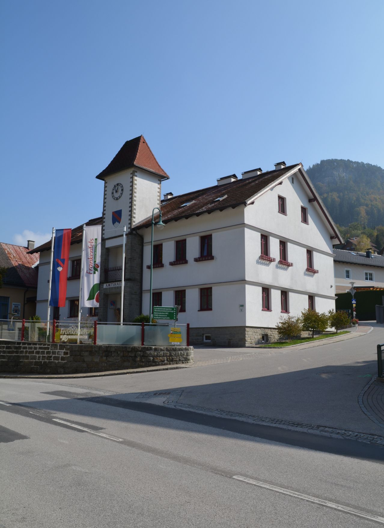Ein großes weißes Gebäude mit einem Uhrturm und mehreren Fahnen befindet sich an einer Ecke. Es hat ein braunes Dach und rot umrahmte Fenster. Ein Berg ist im Hintergrund sichtbar.