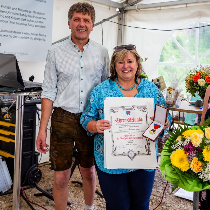 A man and a woman stand in a tent, smiling. The woman holds a certificate and a medal. Behind them is a table with a laptop and a bouquet of flowers.