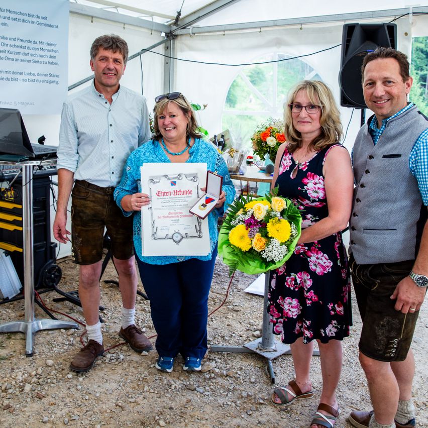 A woman in a blue shirt holds a certificate and a bouquet of flowers, standing between two men in traditional Bavarian attire. They are in a tent with a speaker and a table with flowers.
