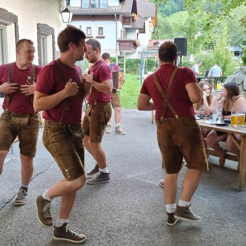 Four men in traditional Bavarian attire, including brown leather shorts and suspenders, are dancing outside a building. In the background, a table with glasses of beer and people sitting is visible.