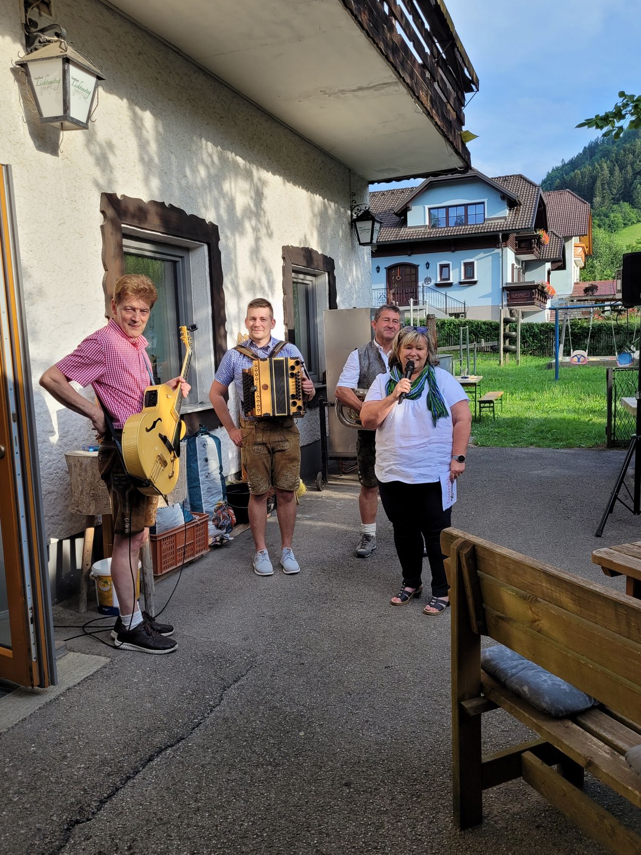 Three people stand outside a house, one holding a guitar, another playing an accordion, and a woman holding a microphone.