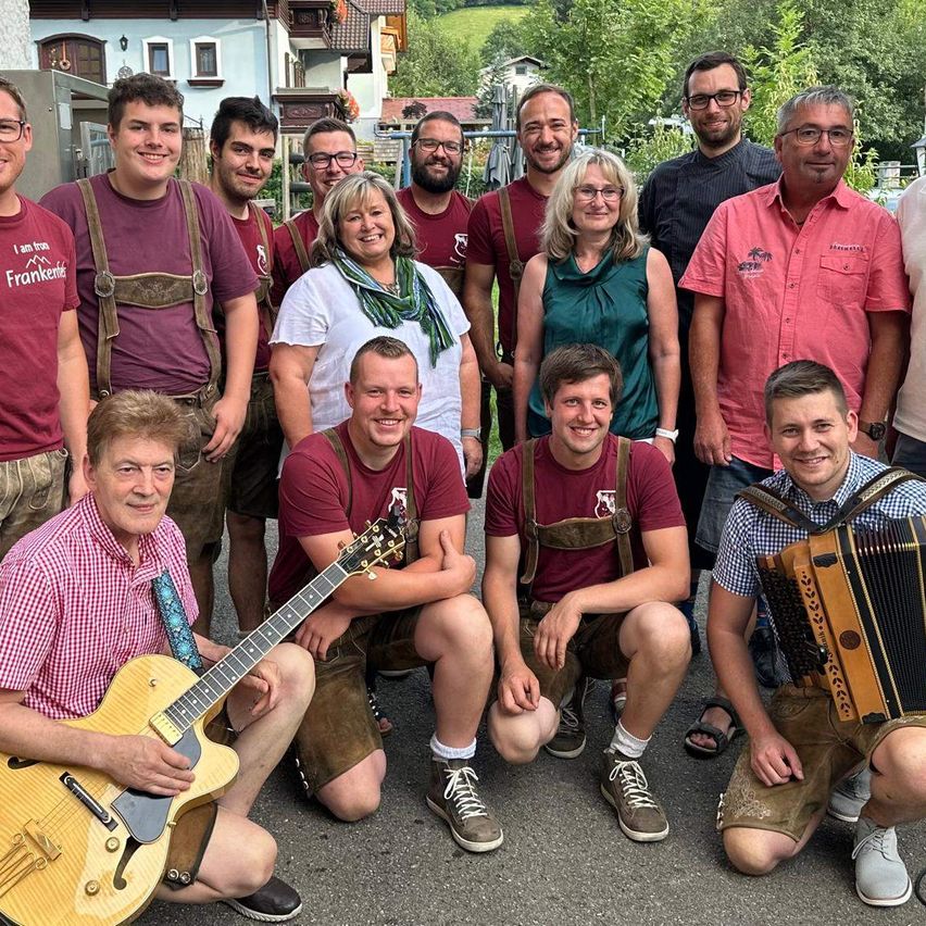 A group of people dressed in traditional Bavarian attire, including lederhosen, are posing for a photo with musical instruments in a scenic outdoor setting.