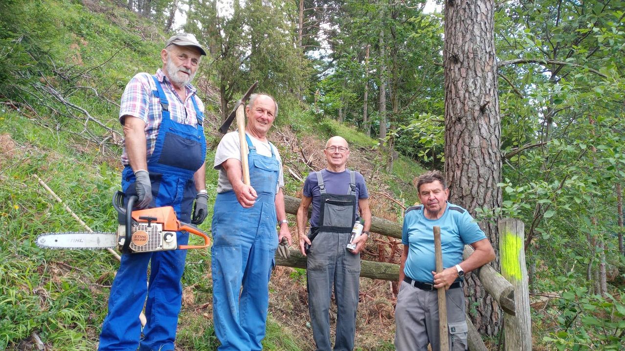 Vier Männer in Latzhosen und Handschuhen stehen in einem Wald und lächeln für ein Foto. Einer hält eine Kettensäge, ein anderer einen Hammer und ein Dritter eine Flasche. Hinter ihnen steht ein Holzzaun.