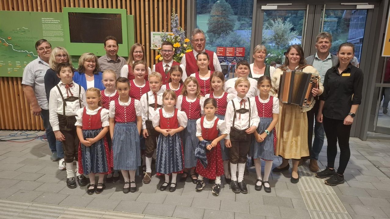 Eine Gruppe von Kindern in traditioneller deutscher Kleidung, zwei Erwachsene vorne, einer hält ein Akkordeon. Sie stehen in einer Reihe mit einem Gebäude im Hintergrund und einer Blumenarrangement.