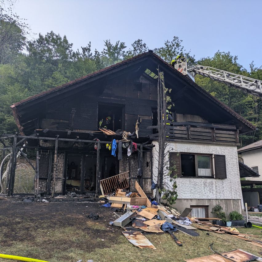 Ein Haus mit beschädigtem Dach und Wänden, Feuerwehrleute sind auf dem Dach und im Haus. Es gibt Trümmer und Rauch in der Umgebung.