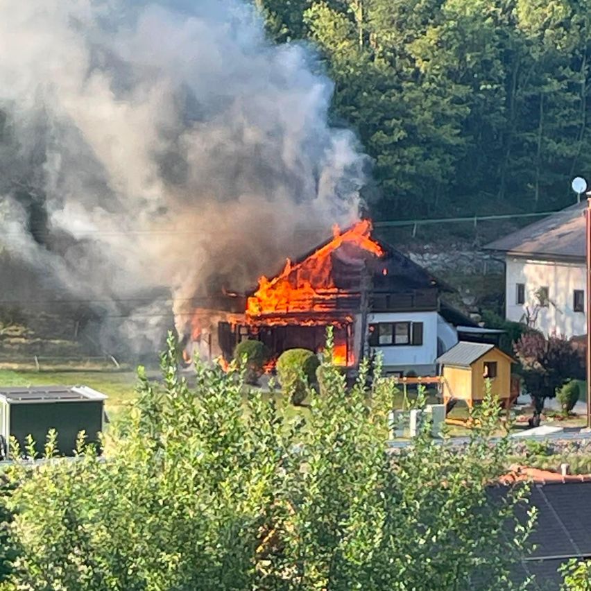 Ein Haus in Flammen mit starkem Rauch und Feuer, umgeben von Bäumen und anderen Häusern in einem ländlichen Gebiet.