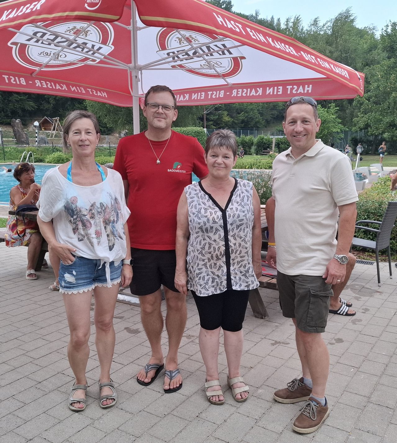 Vier Personen stehen unter einem roten Schirm mit Logo und lächeln für ein Foto. Sie sind lässig gekleidet, und im Hintergrund befindet sich ein Schwimmbad und Grünflächen.