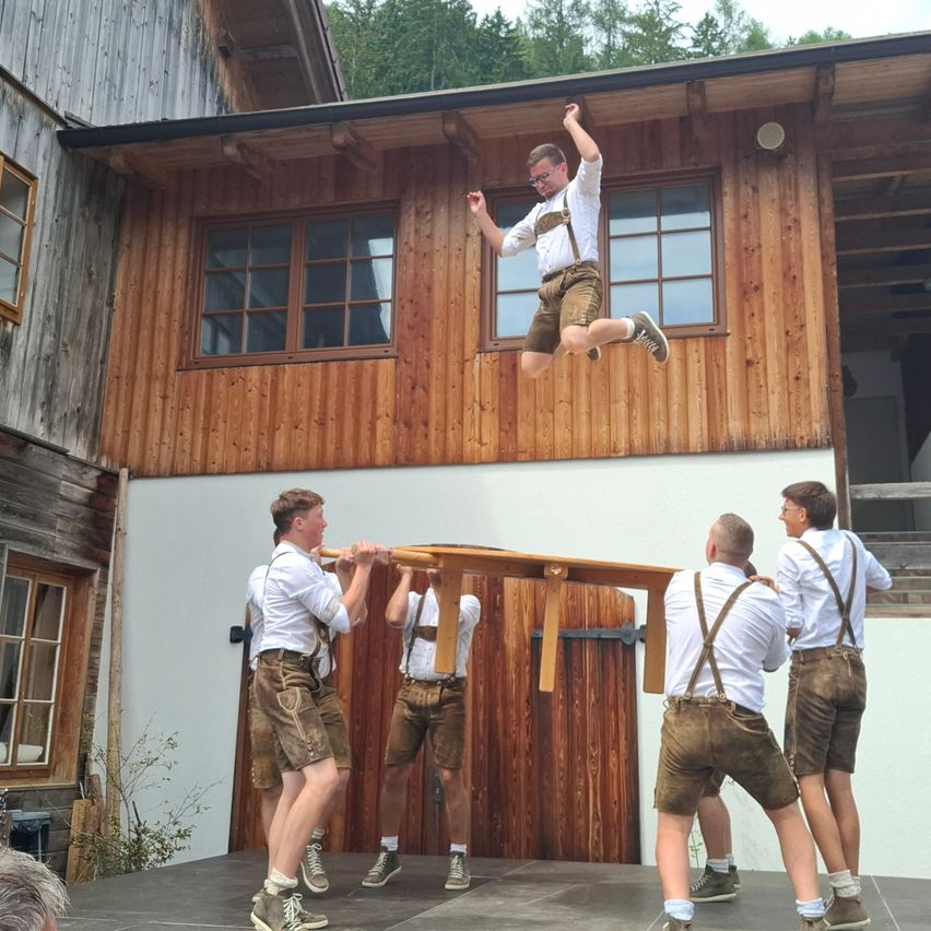 Fünf Männer in traditioneller bayerischer Kleidung heben eine Holzplanke, während einer darüber springt. Sie stehen vor einem Holzhaus.