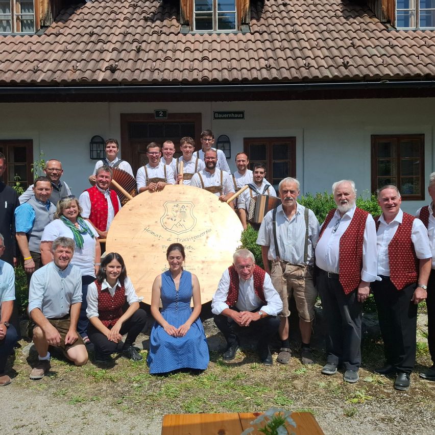 Eine Gruppe von Menschen in traditioneller Kleidung posiert für ein Foto vor einem Gebäude mit dem Schild 'Bauernhaus'. In der Mitte steht ein großer hölzerner Schild mit Text.