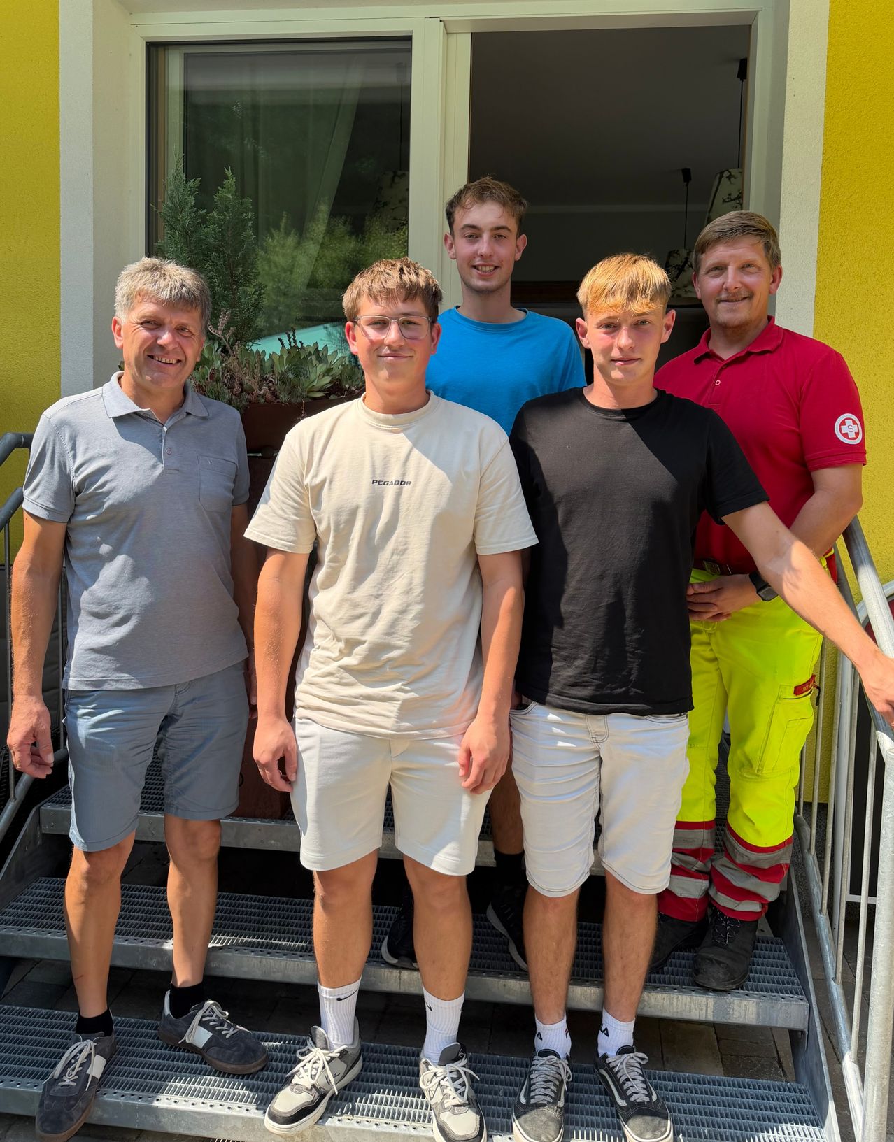 Fünf Männer stehen auf einer Treppe und lächeln für ein Foto. Der Mann ganz links trägt ein graues Shirt und Shorts. Die anderen tragen weiße, schwarze und rote Shirts.