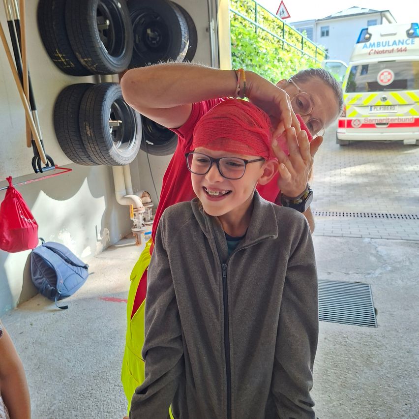 Ein lächelnder Junge mit einem roten Stirnband am Kopf lässt sich von einer Frau in einer Garage die Haare machen. Hinter ihnen hängen Reifen an der Wand, und ein blauer Rucksack liegt auf dem Boden. Ein Krankenwagen ist draußen geparkt.