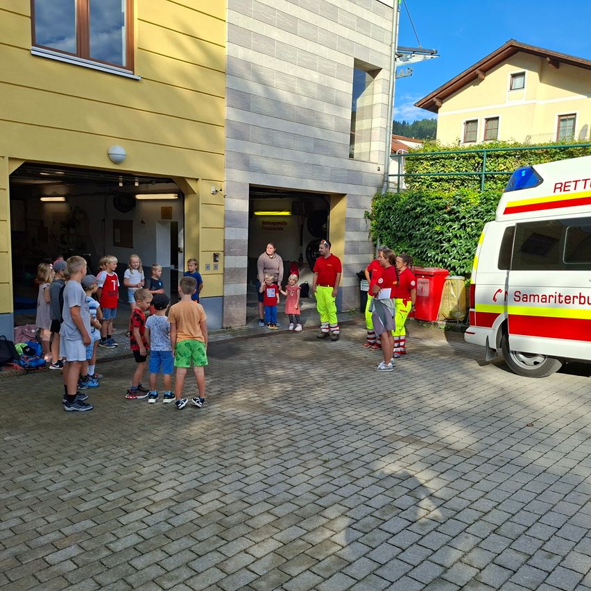 Eine Gruppe von Kindern und Erwachsenen versammelt sich vor einer Feuerwache, mit einem geparkten Krankenwagen in der Nähe. Einige Erwachsene sind in roten Uniformen gekleidet, möglicherweise Feuerwehrleute.