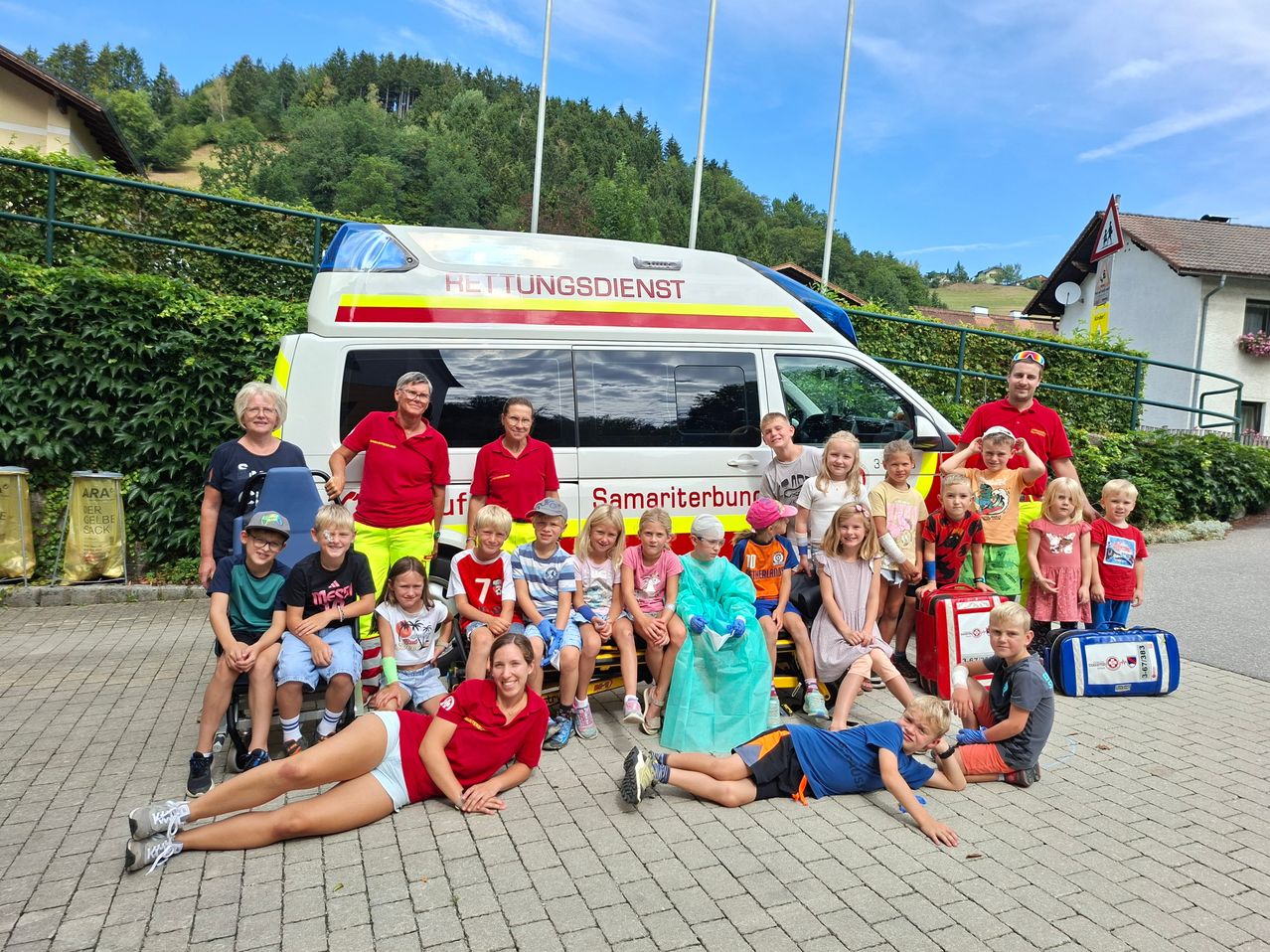 Eine Gruppe von Kindern und Erwachsenen in roten und blauen Uniformen posiert für ein Foto vor einem Krankenwagen. Einige sitzen auf dem Krankenwagen, während andere stehen.