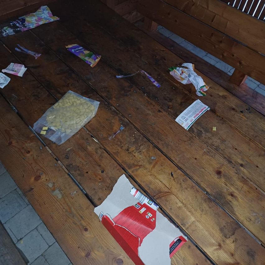 Eine hölzerne Bank mit verschiedenen Lebensmittelverpackungen, Plastiktüten und anderen Gegenständen, die darauf verstreut sind. Einige Gegenstände sind in Plastiktüten und andere auf der Bank. Die Bank hat einige Flecken.