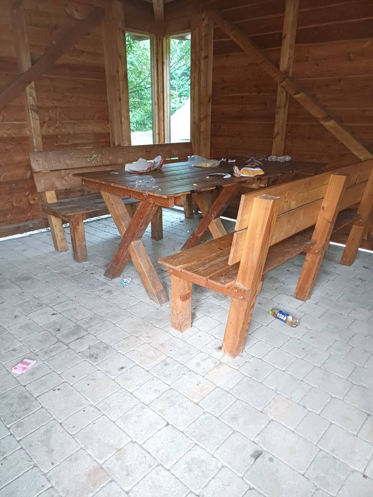 Ein hölzerner Tisch und Bänke sind in einem hölzernen Raum mit Ziegelboden aufgestellt. Es gibt Müll auf dem Tisch und Boden.