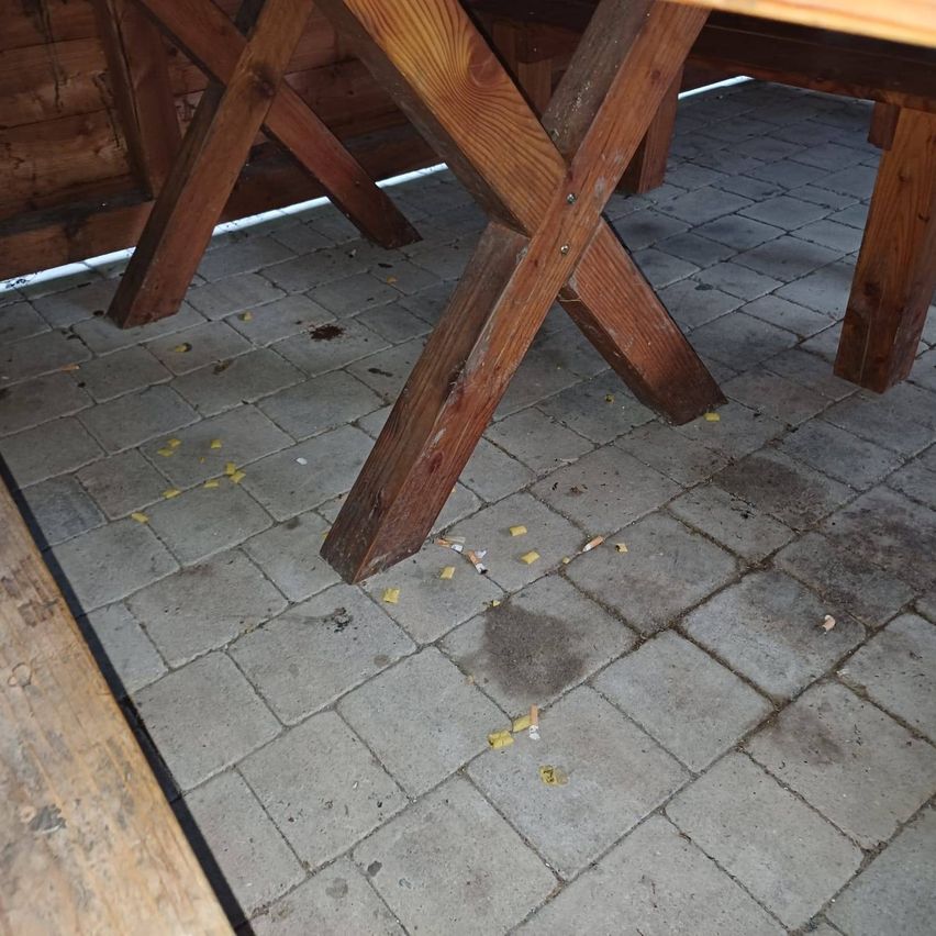 Ein hölzerner Außentisch mit Ziegelboden und verstreuten Zigarettenkippen und gelben Papierstücken.