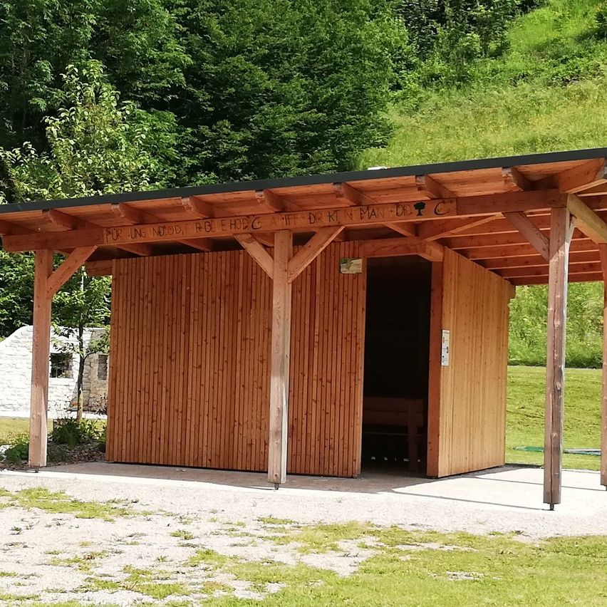 Eine hölzerne Struktur mit einem schwarzen Dach und einer Holzbank im Inneren, umgeben von grünem Gras und Bäumen.