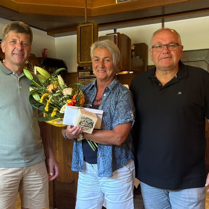 Drei Menschen stehen lächelnd in einem Raum. Eine Frau in der Mitte hält einen Blumenstrauß und eine Schachtel. Der Mann links trägt ein helles Poloshirt, der Mann rechts ein dunkles Poloshirt. Hinter ihnen sind Holzschränke und Regale zu sehen.