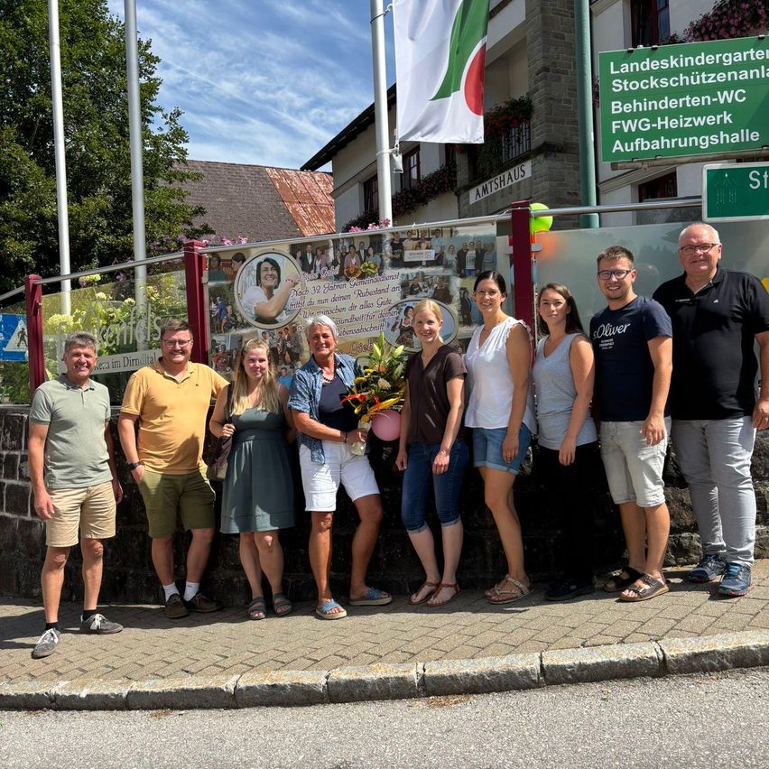 Eine Gruppe von Menschen steht vor einer Wand mit einer Collage von Fotos und einem Schild, das 'Landeskindergarten Stockschützenanlage Behinderten-WC' besagt. Sie lächeln und posieren für ein Foto.