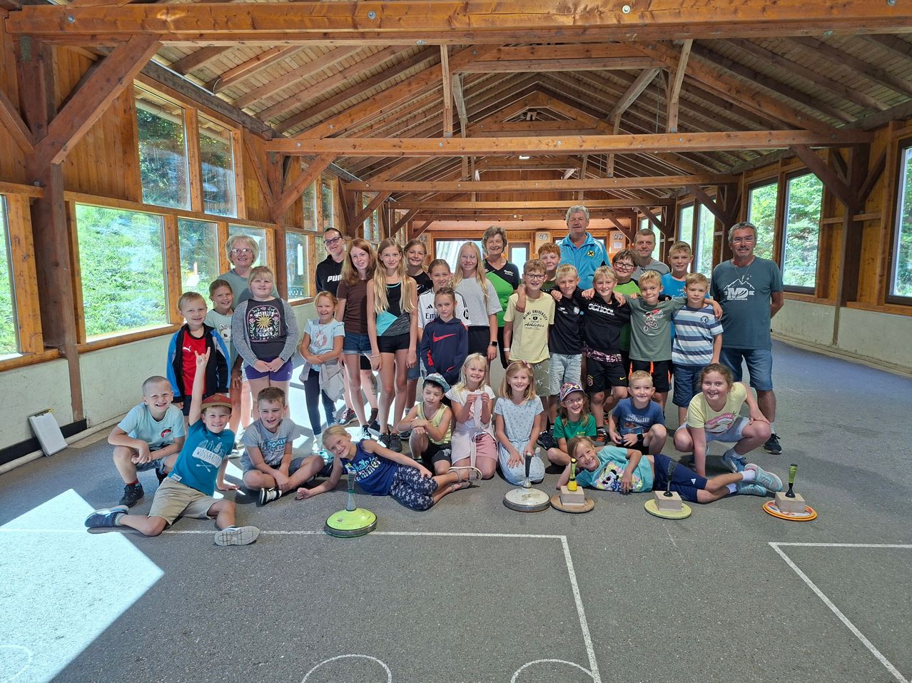 Eine Gruppe von Kindern und Erwachsenen posiert für ein Foto in einem hölzernen Raum mit großen Fenstern und einem Betonboden. Einige Kinder sitzen auf dem Boden, während andere hinter ihnen stehen.